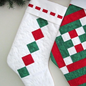 May include: Two Christmas stockings. One is white with red and green diamond-shaped patches. The other stocking has a red, green, and white patchwork design. Both stockings have a white cuff with red accents. Festive holiday decor.