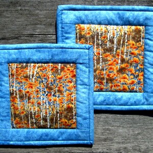 May include: Two blue quilted placemats with a white birch tree and orange leaf pattern in the center.