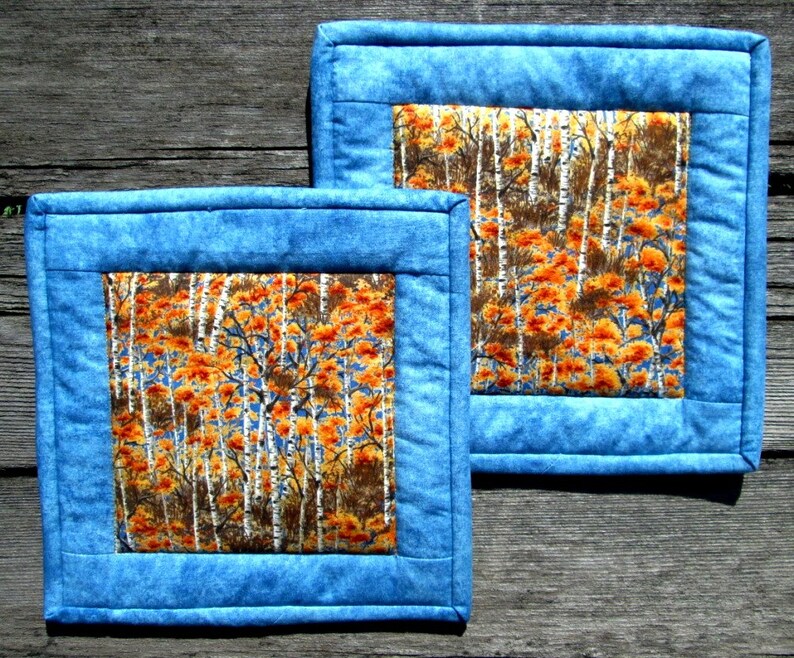 May include: Two blue quilted placemats with a white birch tree and orange leaf pattern in the center.