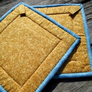 May include: Two quilted pot holders with a blue trim and a gold floral pattern.