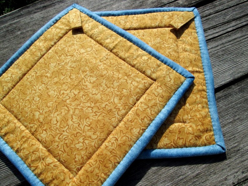 May include: Two quilted pot holders with a blue trim and a gold floral pattern.