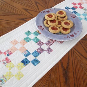May include: A white quilted table runner with a patchwork design in pastel colors, including pink, blue, and yellow squares. A light blue plate with heart accents holds several cookies with jam filling. The runner is on a wooden table.