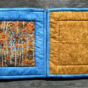 May include: Two blue and yellow quilted pot holders with a fall-themed fabric square in the center of one pot holder.
