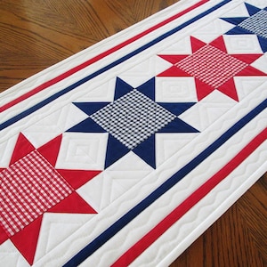 May include: A patriotic table runner featuring a repeating star pattern in red, white, and blue. The stars have a gingham center and are surrounded by red points. The runner is approximately 60 inches long.