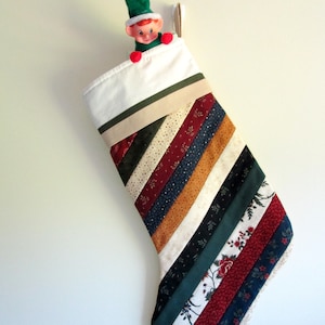 May include: A white Christmas stocking with a green, red, and brown striped pattern. A small elf doll is peeking out of the top of the stocking.