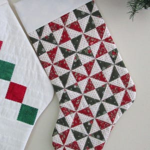 May include: A Christmas stocking with a white cuff and a patchwork design of red, green, and white triangles. The stocking is decorated with a festive pattern and is likely used for holiday gift-giving.