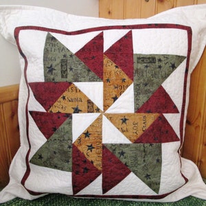 May include: A white pillow with a red, green, and brown patchwork design featuring a starburst pattern. The fabric has a variety of Christmas-themed words and stars printed on it.