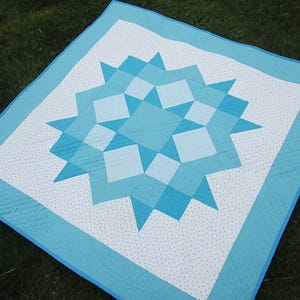 May include: A square quilt featuring a central star pattern in shades of light and medium blue. The design includes white fabric sections with small blue dots, surrounded by a solid light blue border. The fabric displays visible stitching.