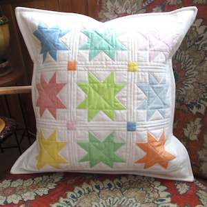 May include: Square white pillow with a patchwork design featuring colorful star shapes in pastel shades of blue, green, pink, yellow, and orange. The pillow is quilted with a white border and a white background.