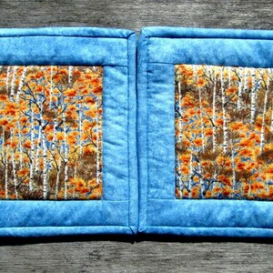 May include: Two blue quilted pot holders with a square inner panel featuring a white birch tree and orange leaf pattern.