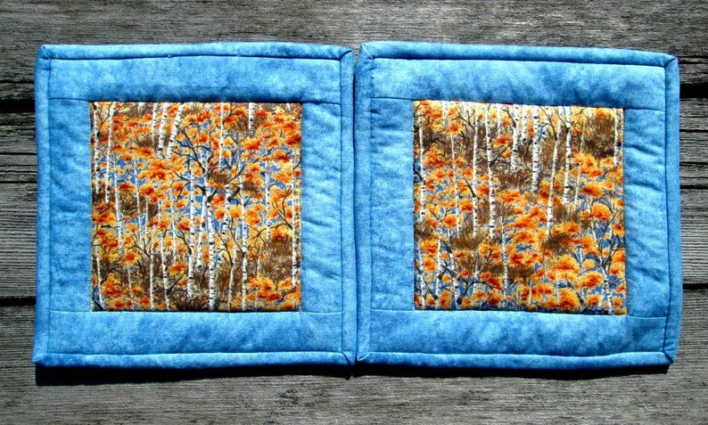 May include: Two blue quilted pot holders with a square inner panel featuring a white birch tree and orange leaf pattern.