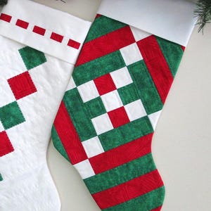 May include: Two Christmas stockings with white cuffs and quilted designs. One stocking features a pattern of red squares and green diamonds on a white background. The other stocking has a patchwork design of red, green, and white squares and stripes.