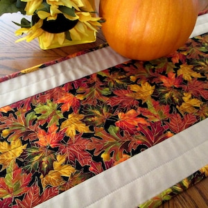 Table runner of scattered colorful autumn leaves edged with a fine line of gold on black, bordered on each side with tan. 41” x 10 1/2”
