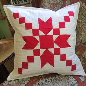 May include: A square throw pillow featuring a white fabric base with a prominent red geometric star pattern centered on the front. The design is constructed with quilted stitching, creating a grid texture across the surface of the pillow cover.