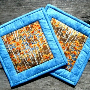 May include: Two blue quilted pot holders with a fall-themed fabric center featuring a pattern of orange and brown leaves and white birch trees.