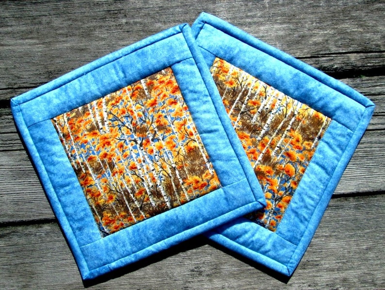May include: Two blue quilted pot holders with a fall-themed fabric center featuring a pattern of orange and brown leaves and white birch trees.