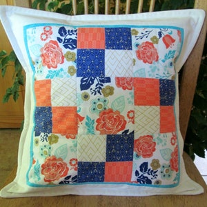 May include: A white pillow with a turquoise border and a patchwork design. The patchwork is made up of squares of coral, navy blue, and gold fabric with floral patterns.