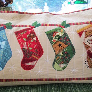 May include: A rectangular quilted throw pillow in beige featuring four appliqued Christmas stockings in blue, red, green, and cream. The stockings hang from a horizontal plaid strip with green holly leaves and red berries. White star stitching accents.