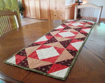 May include: A quilted table runner with a geometric pattern in shades of red, brown, and beige. The runner features a central design of diamond shapes and triangles, with a green border. The runner is placed on a round wooden table.
