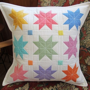 May include: Square white pillow with a patchwork design featuring nine colorful star patterns in yellow, pink, blue, turquoise, green, purple, and orange. The stars are set against a white background with quilted lines.
