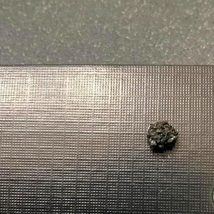 May include: A small, dark gray, irregularly shaped rock on a silver textured surface.