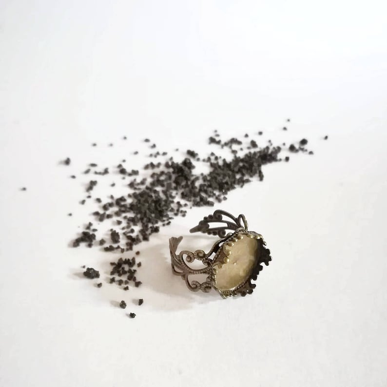 May include: A gold-toned ring with a round, clear glass bezel setting. The ring is on a white surface with scattered black specks.