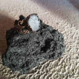 May include: A silver ring with a white stone set in a bronze setting. The ring is sitting on a dark grey rock.