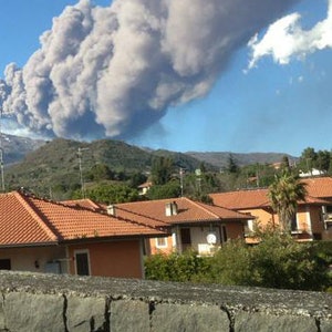 May include: A large plume of smoke rises from a volcano in the distance, with houses and trees in the foreground.
