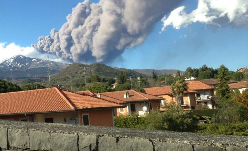 May include: A large plume of smoke rises from a volcano in the distance, with houses and trees in the foreground.