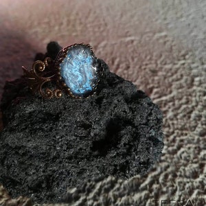 May include: A ring with a blue and black resin cabochon set in a bronze setting. The ring is on a black rock.