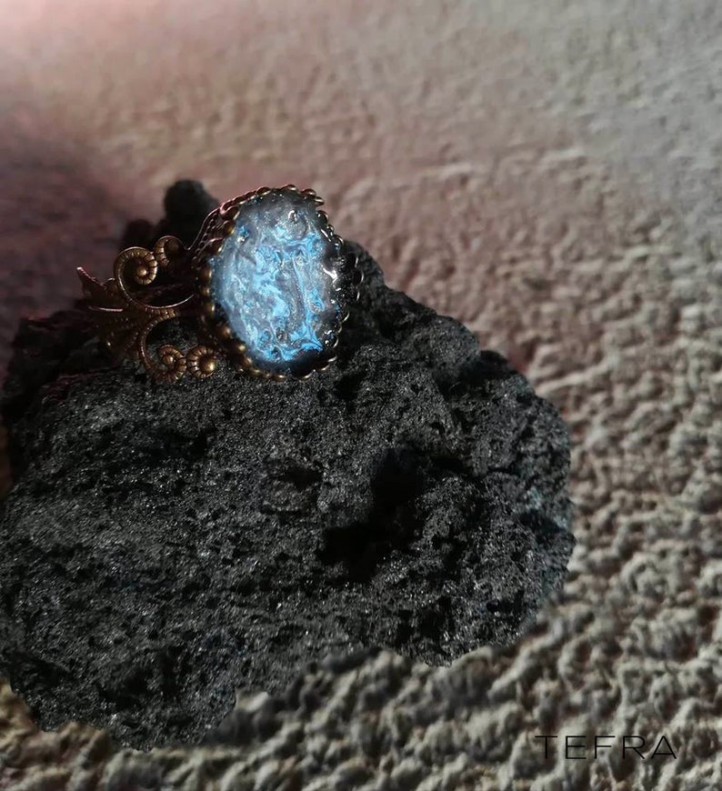 May include: A ring with a blue and black resin cabochon set in a bronze setting. The ring is on a black rock.