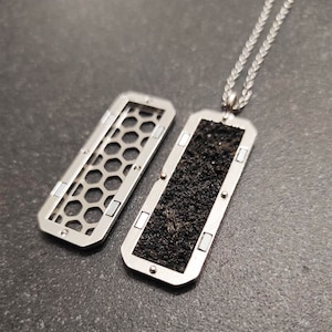 May include: A silver necklace with a rectangular pendant. The pendant has a black, textured surface on one side and a honeycomb pattern on the other side.