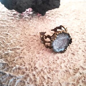 May include: A silver ring with a clear, textured stone set in a decorative, ornate frame. The ring is on a textured, light brown surface.