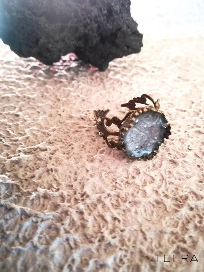 May include: A silver ring with a clear, textured stone set in a decorative, ornate frame. The ring is on a textured, light brown surface.
