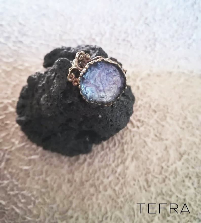 May include: A ring featuring a round, purple and white resin cabochon set in a gold-toned filigree setting. The ring is resting on a dark grey rock.