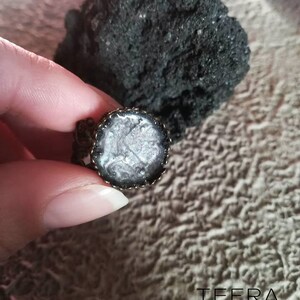 May include: A silver and black ring with a round, clear resin inlay. The resin has a swirling, marbled pattern.