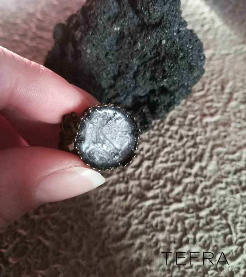 May include: A silver and black ring with a round, clear resin inlay. The resin has a swirling, marbled pattern.