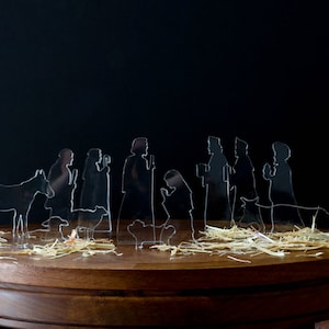 May include: A set of clear acrylic Christmas nativity figures on a wooden table with straw. The figures include Mary, Joseph, the baby Jesus, a donkey, a sheep, and three wise men.