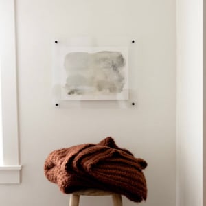 May include: A brown knitted throw blanket draped over a wooden stool in front of a white wall with a framed abstract watercolor painting.
