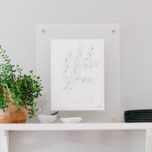 May include: A black and white line drawing of a dove with an olive branch in its beak, framed in a clear acrylic frame. The artwork is hanging on a white wall above a white shelf.