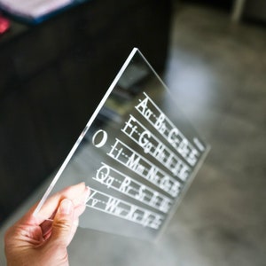 May include: A clear acrylic sheet with a hole in the top left corner. The sheet has white lines and letters printed on it, spelling out the alphabet in rows. The letters are in a cursive font.