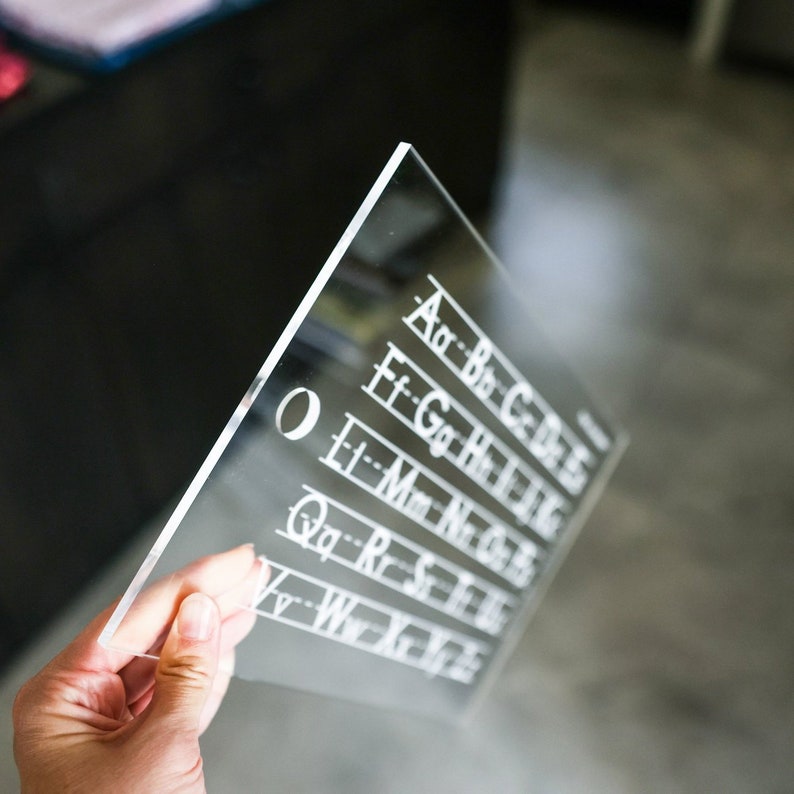 May include: A clear acrylic sheet with a hole in the top left corner. The sheet has white lines and letters printed on it, spelling out the alphabet in rows. The letters are in a cursive font.