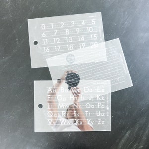 May include: Two clear plastic stencils with white numbers and letters. The top stencil has numbers 0-20. The bottom stencil has the alphabet.