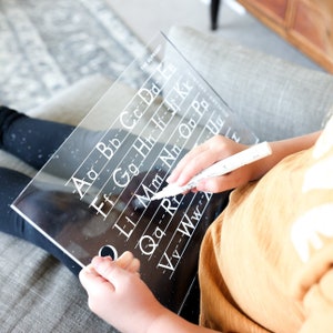 May include: A clear acrylic dry-erase board with the alphabet printed on it. The board is being used by a child to practice writing the letters.