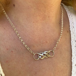 May include: A silver-colored necklace with a chain and a central pendant. The pendant features an intertwined design of three connected loops. The necklace is worn around the neck, showcasing its simple yet elegant design.