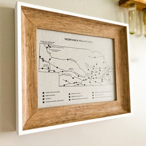 May include: A framed map of Nebraska railroads with a white mat and a brown wood frame. The map shows the different railroad lines and cities in the state. The title of the map is "Nebraska Railroads".