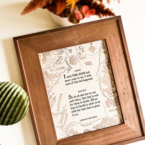 Puede incluir: Un marco de madera marrón que contiene una impresión con un mapa y citas de "El Señor de los Anillos". El fondo es beige con un mapa detallado y texto del libro.