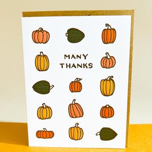 May include: A greeting card with a white front featuring illustrated pumpkins in orange, yellow, and green. The words "MANY THANKS" are printed in the centre. The card is set against a yellow background.