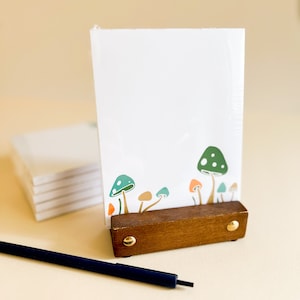 May include: A white notepad with a mushroom illustration on the front. The notepad is in a wooden stand with gold accents. A black pen is lying on a beige surface.