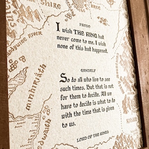 Può includere: Stampa incorniciata con una mappa della Terra di Mezzo e citazioni dal Signore degli Anelli. La mappa è in toni seppia, con testi di Frodo e Gandalf. La cornice in legno aggiunge un tocco rustico.
